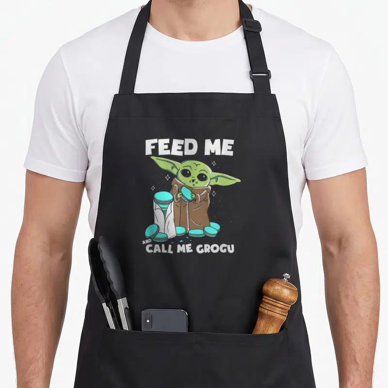 Tablier de cuisine noir avec illustration de Grogu tenant un macaron turquoise et le texte Feed Me and Call Me Grogu en blanc.