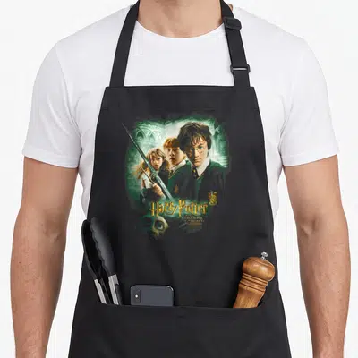 Tablier de cuisine noir avec Harry Potter, Hermione et Ron tenant une épée devant un décor vert sombre et gothique.