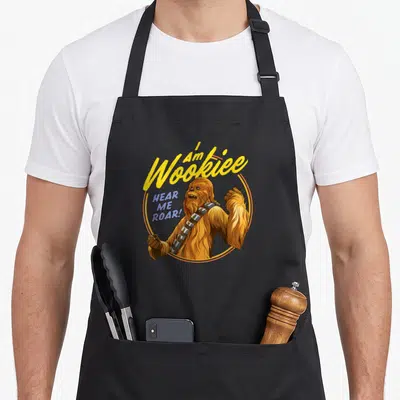 Tablier de cuisine noir avec une illustration colorée de Chewbacca et du texte jaune et bleu sur fond uni.