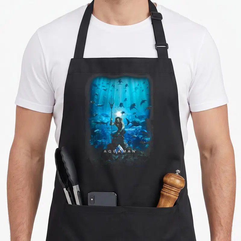 Tablier de cuisine noir avec illustration d'Aquaman tenant un trident au milieu de requins sous une lumière bleue.