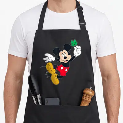 Tablier de cuisine noir avec Mickey Mouse sautant et tenant un trèfle à quatre feuilles vert dans sa main gantée de blanc.