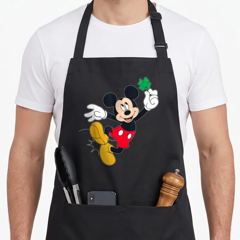 Tablier de cuisine noir avec Mickey Mouse sautant et tenant un trèfle à quatre feuilles vert dans sa main gantée de blanc.