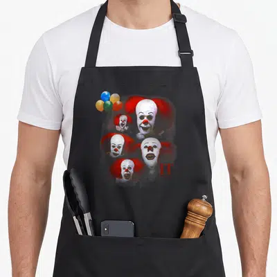 Tablier de cuisine noir avec cinq visages du clown Pennywise, des ballons multicolores et le texte IT The Movie imprimé en rouge.