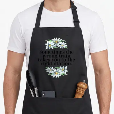 Tablier de cuisine noir avec une citation en anglais entourée de bouquets de fleurs blanches aux centres jaunes.