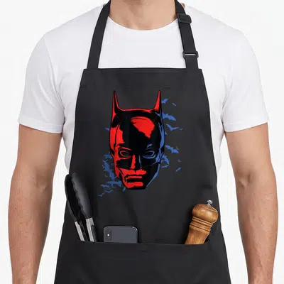 Tablier de cuisine noir avec le masque de Batman en rouge vif et des chauves-souris bleues en arrière-plan.