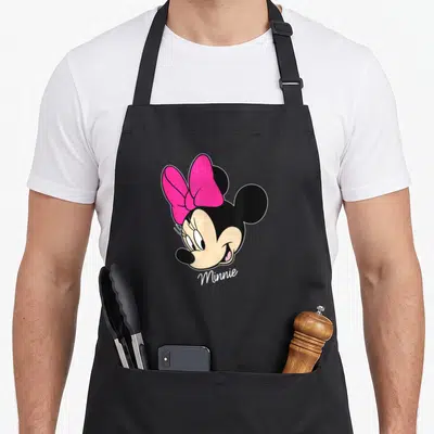 Tablier de cuisine noir avec le visage souriant de Minnie et son grand ruban rose vif.