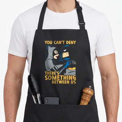Tablier de cuisine noir avec illustration vintage de Batman et Catwoman et texte jaune You Can't Deny There's Something Between Us.