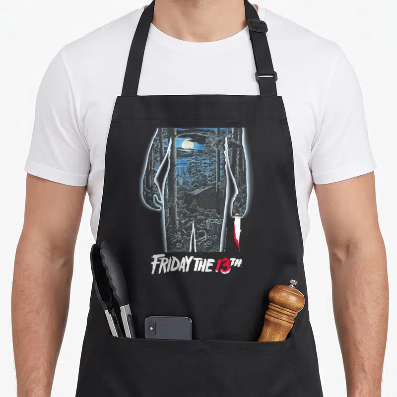 Tablier de cuisine noir avec illustration d'une cabane en forêt, lune pleine et couteau ensanglanté sous le titre Friday the 13th.