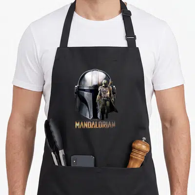 Tablier de cuisine noir avec un grand casque de Mandolorien et une version miniature du personnage debout devant le logo doré.