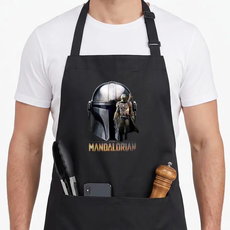 Tablier de cuisine noir avec un grand casque de Mandolorien et une version miniature du personnage debout devant le logo doré.