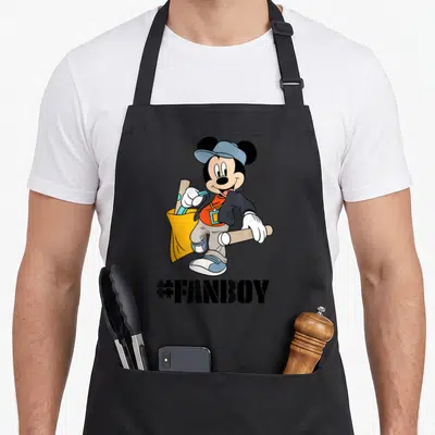 Tablier de cuisine noir avec Mickey Mouse en tenue décontractée tenant des bâtons et le texte #FANBOY imprimé en dessous.