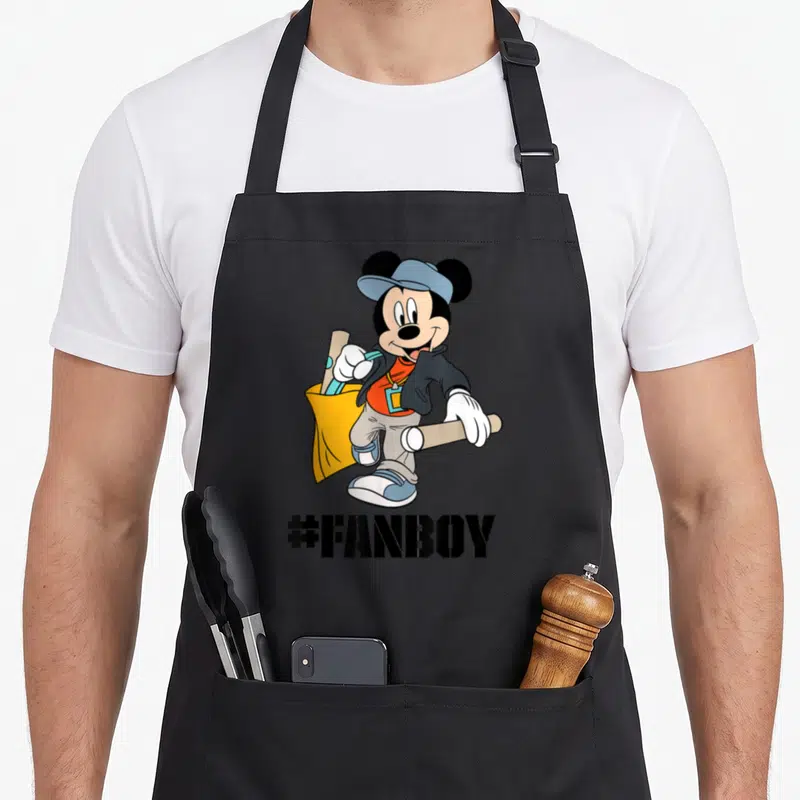 Tablier de cuisine noir avec Mickey Mouse en tenue décontractée tenant des bâtons et le texte #FANBOY imprimé en dessous.