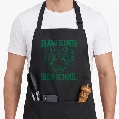 Tablier de cuisine noir avec le visage d'un tige et l'inscription Hawkins High School 1986 en vert délavé.