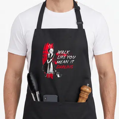 Tablier de cuisine noir avec illustration de Cruella et texte Walk Like You Mean It Darling en blanc et rouge.