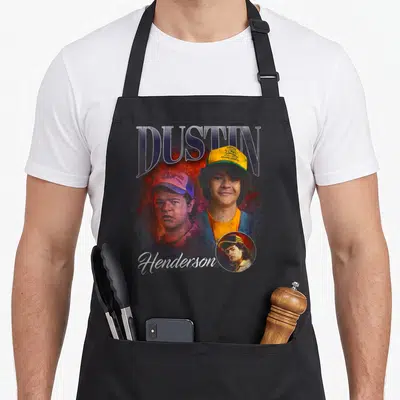 Tablier de cuisine noir avec illustration de Dustin Henderson et son casque jaune typique.