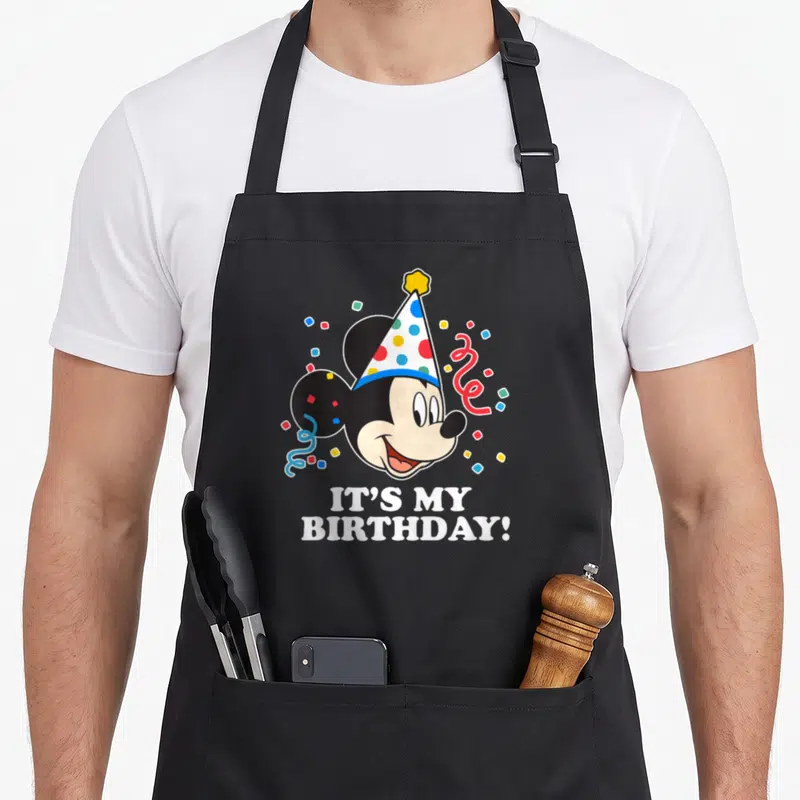 Tablier de cuisine noir avec tête de Mickey Mouse au chapeau d'anniversaire et confettis colorés.