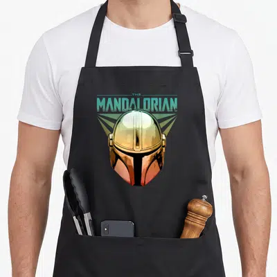 Tablier de cuisine noir avec illustration du casque doré du Mandalorian et texte turquoise The Mandalorian au-dessus.