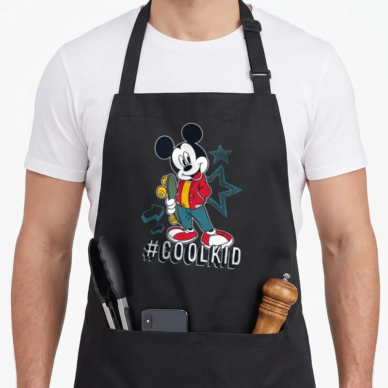 Tablier de cuisine noir avec Mickey Mouse en tenue urbaine tenant un skateboard et le texte #COOLKID imprimé en bas.