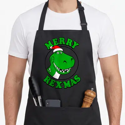 Tablier de cuisine noir avec Rex le dinosaure vert portant un chapeau de Père Noël et l'inscription Merry Rex-Mas.