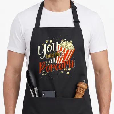 Tablier de cuisine noir avec illustration de boîte de popcorn rouge et blanche et texte You Had Me At Popcorn en typographie stylisée.