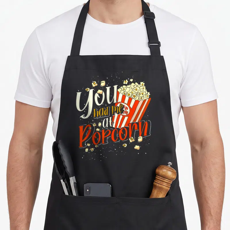 Tablier de cuisine noir avec illustration de boîte de popcorn rouge et blanche et texte You Had Me At Popcorn en typographie stylisée.