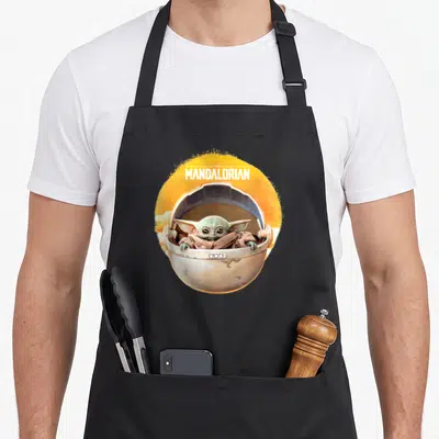 Tablier de cuisine noir avec Grogu dans son pod volant et le logo The Mandalorian sur fond orange circulaire.