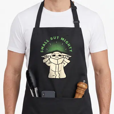 Tablier de cuisine noir avec illustration mignonne de Grogu levant les bras sous le texte Small But Mighty vert.