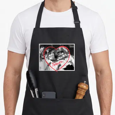 Tablier de cuisine noir avec une scène en noir et blanc de deux personnages dans un grand cœur rouge tracé à la main.