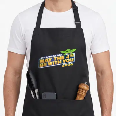 Tablier de cuisine noir avec logo May The 4th Be With You 2026 et petit personnage vert aux grandes oreilles posé sur le texte.