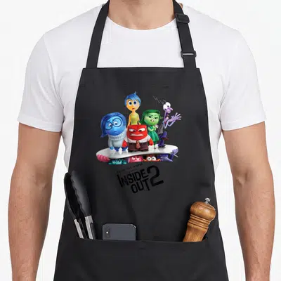 Tablier de cuisine noir avec les personnages d'Inside Out 2 posés sur une surface blanche au-dessus du titre du film.