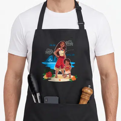Tablier de cuisine noir avec Moana et une enfant debout sur une plage tropicale entourée de fleurs et d'inscriptions.