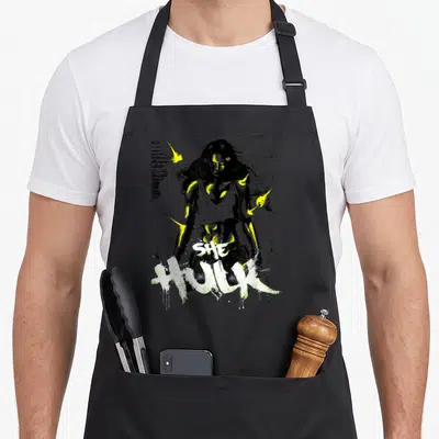 Tablier de cuisine noir avec illustration She-Hulk style graffiti vert jaune et texte She Hulk en lettres blanches éclabouillées.