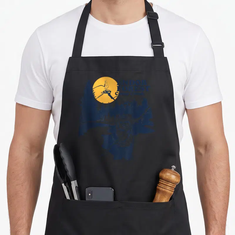Tablier de cuisine noir avec illustration d'un Ewok tenant une lance sous un soleil orange et des arbres bleus.