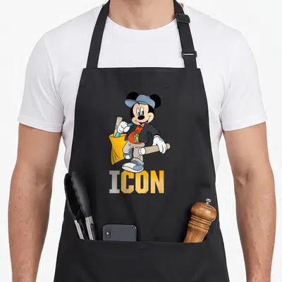 Tablier de cuisine noir avec Mickey Mouse en tenue streetwear tenant un sac jaune et le mot ICON imprimé en dessous.