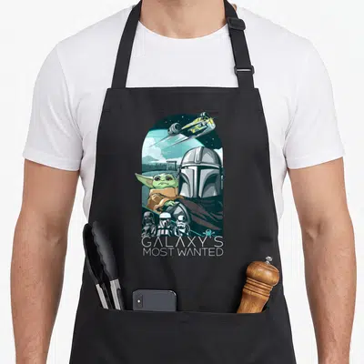 Tablier de cuisine noir avec illustration du Mandalorian, Grogu et un vaisseau spatial sur fond de planète.