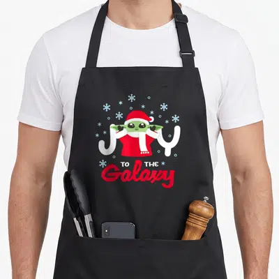 Tablier de cuisine noir avec illustration de Grogu en costume rouge et bonnet de Père Noël sous des flocons de neige.