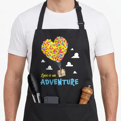 Tablier de cuisine noir avec une maison transportée par un cœur de ballons multicolores et des nuages blancs.