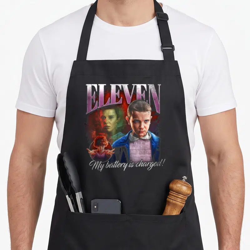 Tablier de cuisine noir avec portrait d'Eleven et texte My battery is charged en bas.