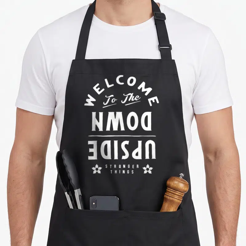 Tablier de cuisine noir avec inscription blanche Welcome to the Upside Down Stranger Things et motifs floraux stylisés.