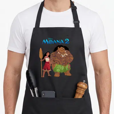 Tablier de cuisine noir avec Moana tenant une rame et Maui aux tatouages visibles sous le logo Moana 2.