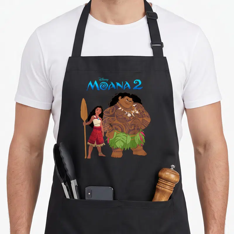 Tablier de cuisine noir avec Moana tenant une rame et Maui aux tatouages visibles sous le logo Moana 2.