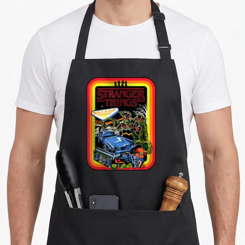 Tablier de cuisine noir avec illustration Stranger Things Arcade, voiture bleue et monstres verts encadrés de couleurs vives.