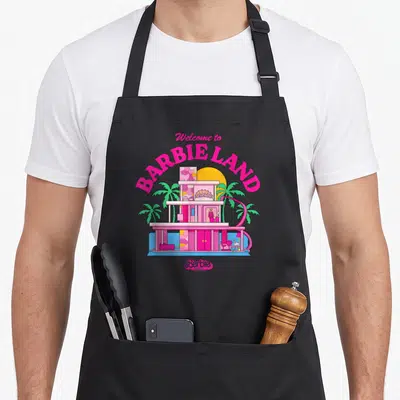 Tablier de cuisine noir avec illustration colorée d'une maison rose et texte Bienvenue à Barbie Land.