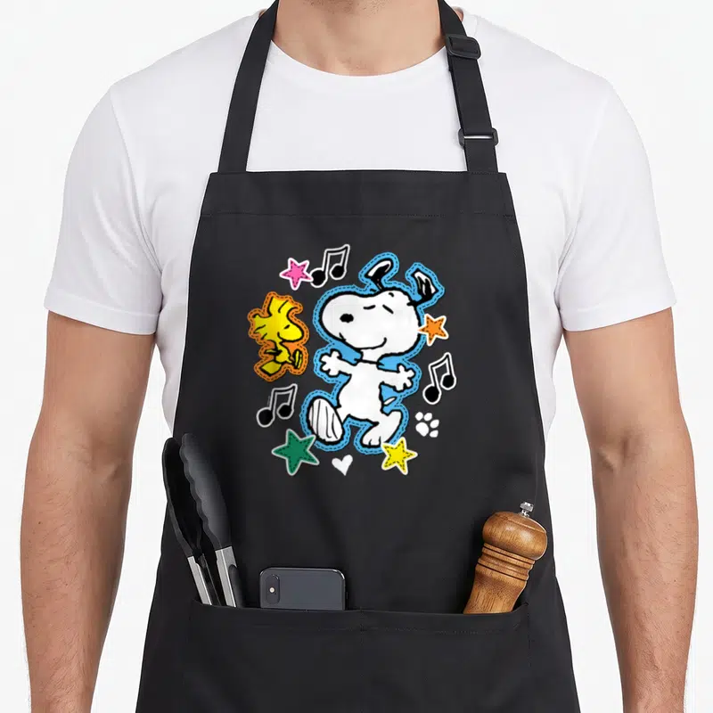 Tablier de cuisine noir avec Snoopy et Woodstock en patchs colorés entourés de notes de musique et d'étoiles.