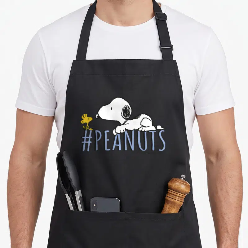 Tablier de cuisine noir avec Snoopy, Woodstock et le texte #PEANUTS en lettres bleues stylisées.