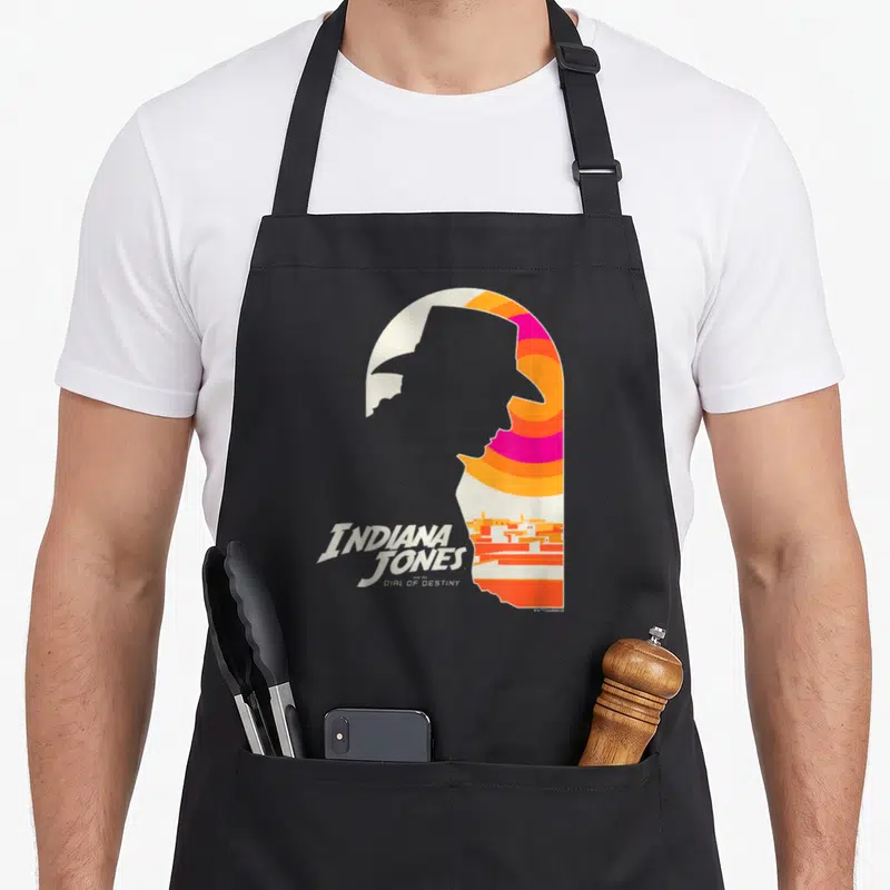 Tablier de cuisine noir avec silhouette stylisée Indiana Jones et texte Dial of Destiny en couleurs vives sur fond uni.