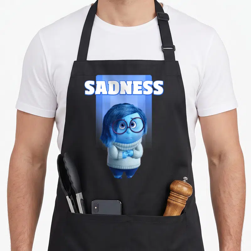 Tablier de cuisine noir avec l'illustration de Sadness en train de se frotter les mains et le texte SADNESS au-dessus.