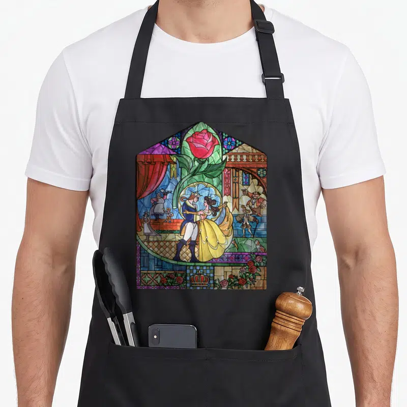 Tablier de cuisine noir avec un motif vitrail coloré montrant Belle et le Prince dansant sous une grande rose rouge.