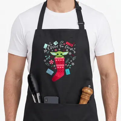Tablier de cuisine noir avec Grogu dans une chaussette de Noël rouge et des cadeaux colorés.
