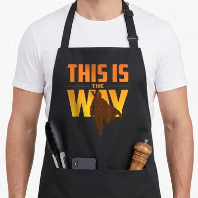 Tablier de cuisine noir avec texte This Is The Way et silhouette du Mandalorian en orange et jaune.